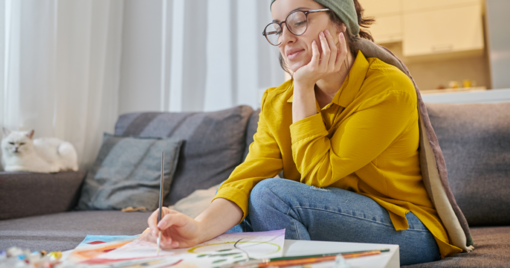 Frau malt konzentriert auf dem Sofa, Pinsel und Farben vor sich
