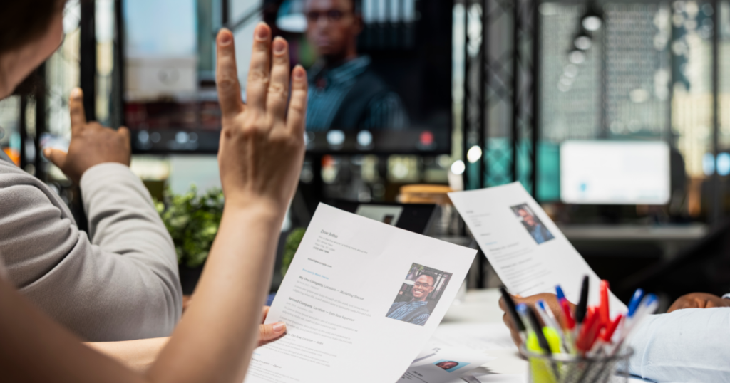 Zwei Personen im Buero sichten Lebenslaeufe, ein Kandidat ist im Videocall zugeschaltet