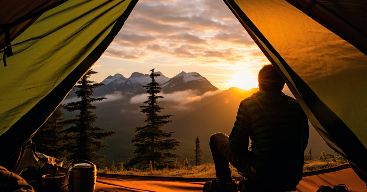 Blick aus einem Zelt auf Berge bei Sonnenaufgang, eine Person sitzt im Gegenlicht