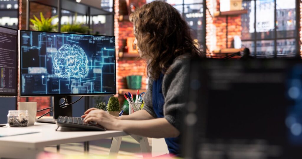 Frau sitzt am Schreibtisch, auf dem Monitor eine Visualisierung kuenstlicher Intelligenz