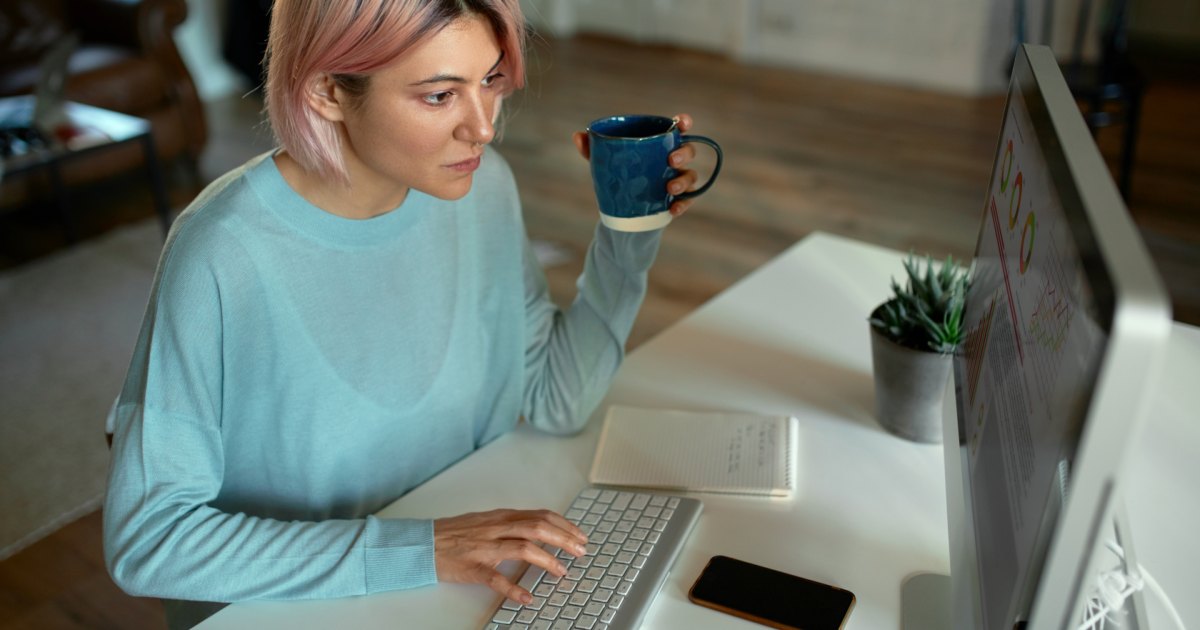 Frau sitzt mit Kaffeetasse am Schreibtisch und arbeitet konzentriert am Computer
