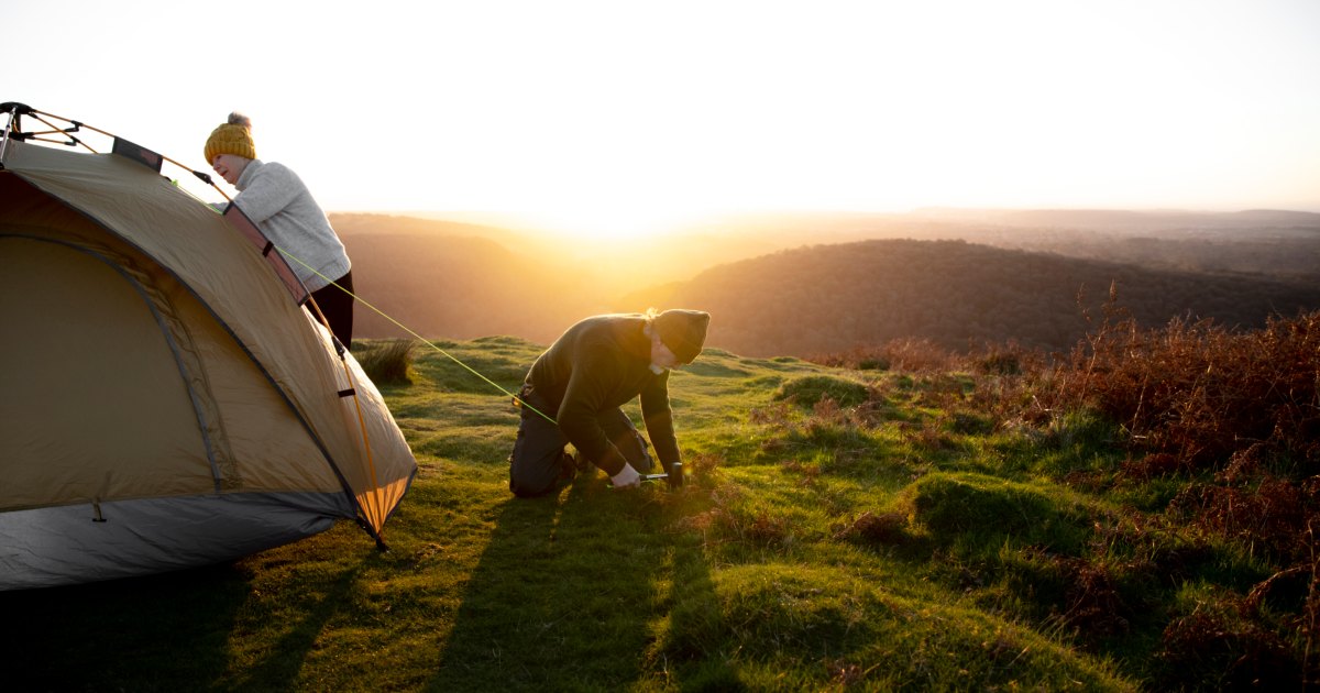 Zwei Personen bauen gemeinsam bei Sonnenaufgang ein Zelt auf einer Anhöhe auf